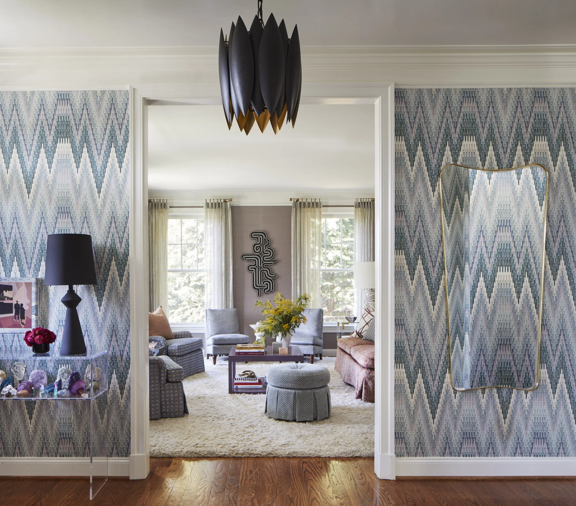 Light-filled sitting room with blue armchairs, a circular ottoman, geometric chandelier, abstract artwork, and layered furnishings.