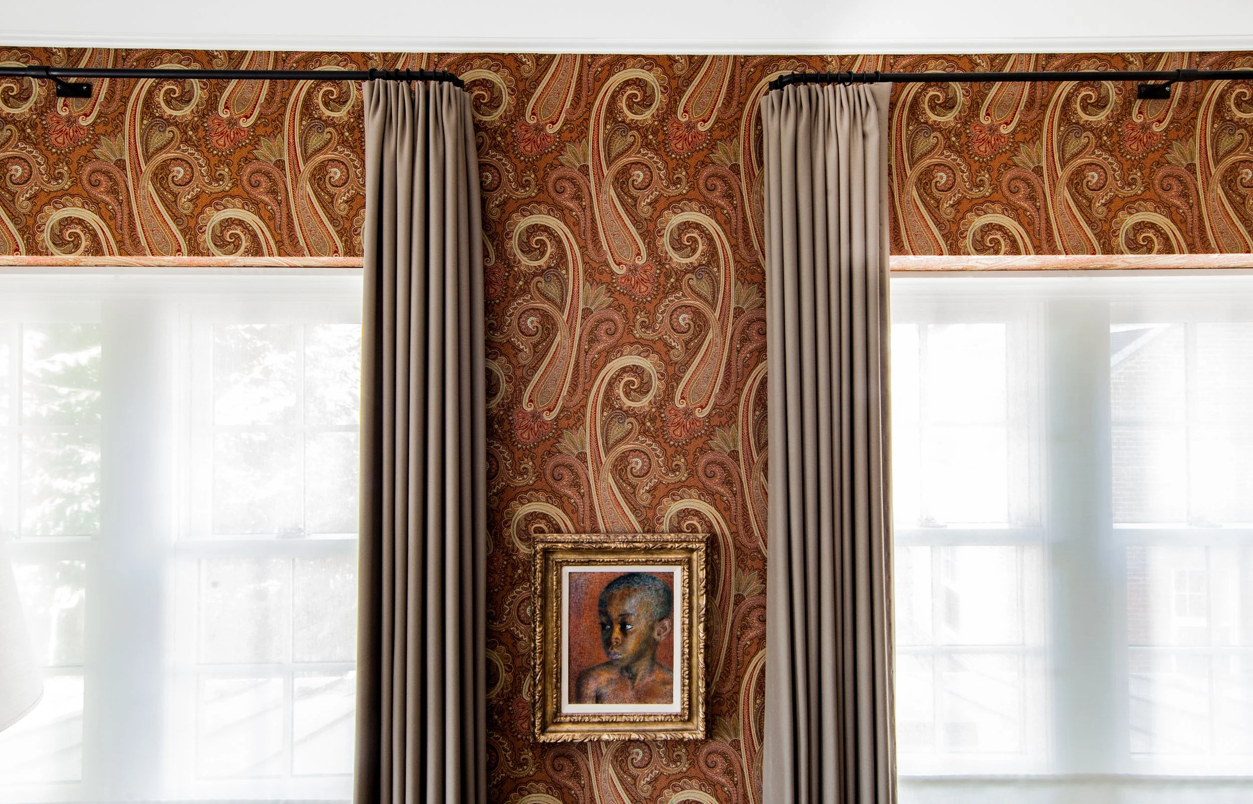 Framed portrait of a child displayed between two windows with beige curtains against paisley wallpaper.