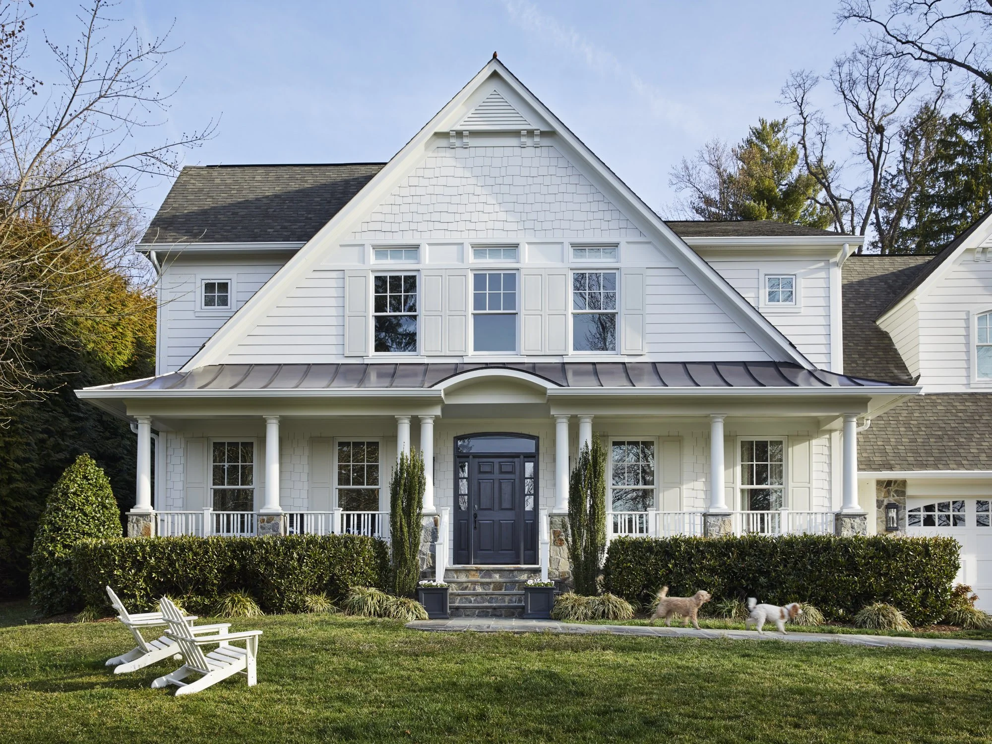 White gabled home with a columned front porch, dark entry door, Adirondack chairs, and mature landscaping.