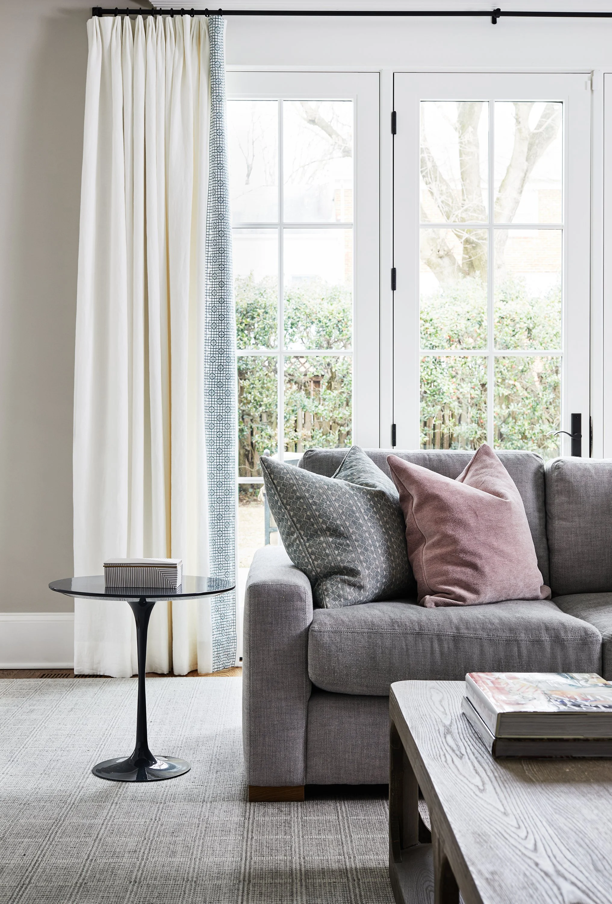 Living room with gray sofa, blush and patterned pillows, black side table, white curtains, and large glass doors.