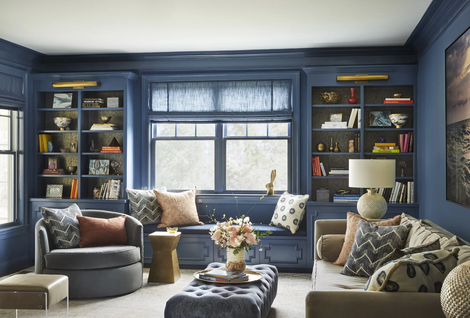 Blue library with custom book-filled shelving and a built-in bench, sculptural round chair, beige sofa, tufted ottoman, and patterned pillows.