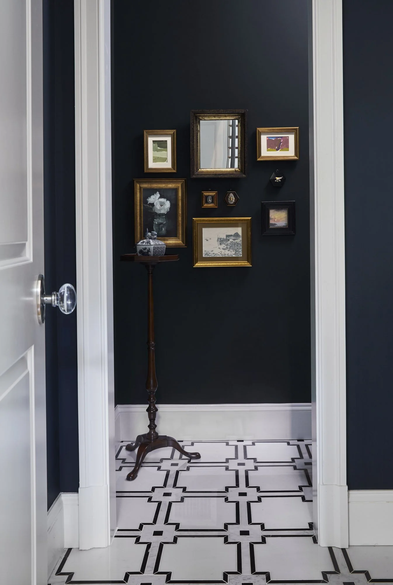 View from a dark hallway into the powder room, with framed artwork, patterned floor tile.
