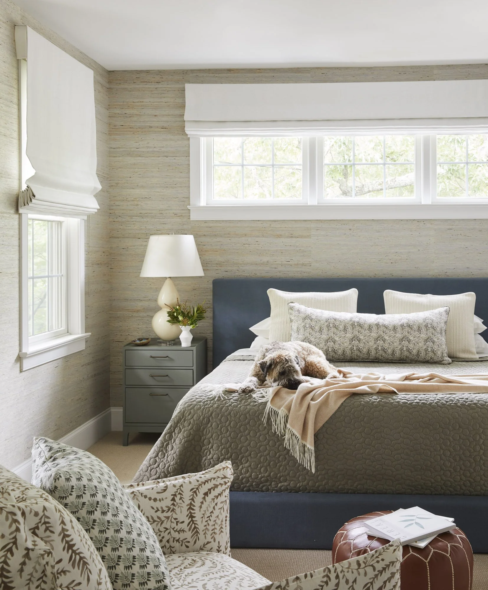 Cozy coastal bedroom with a dog resting on the bed, decorative pillows, a bedside lamp, and a woven pouf.