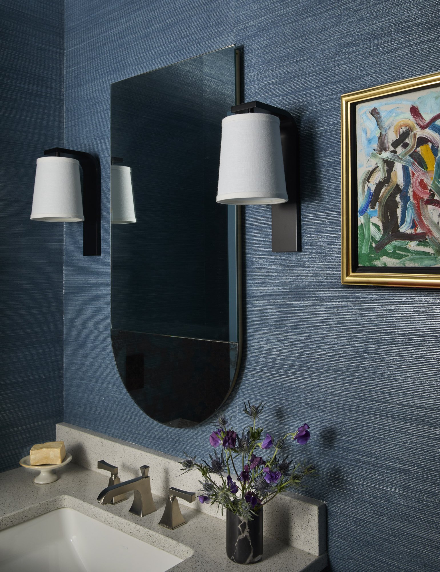 Powder room with textured navy walls, contemporary sconces, a frameless mirror, purple flowers, and colorful abstract art.
