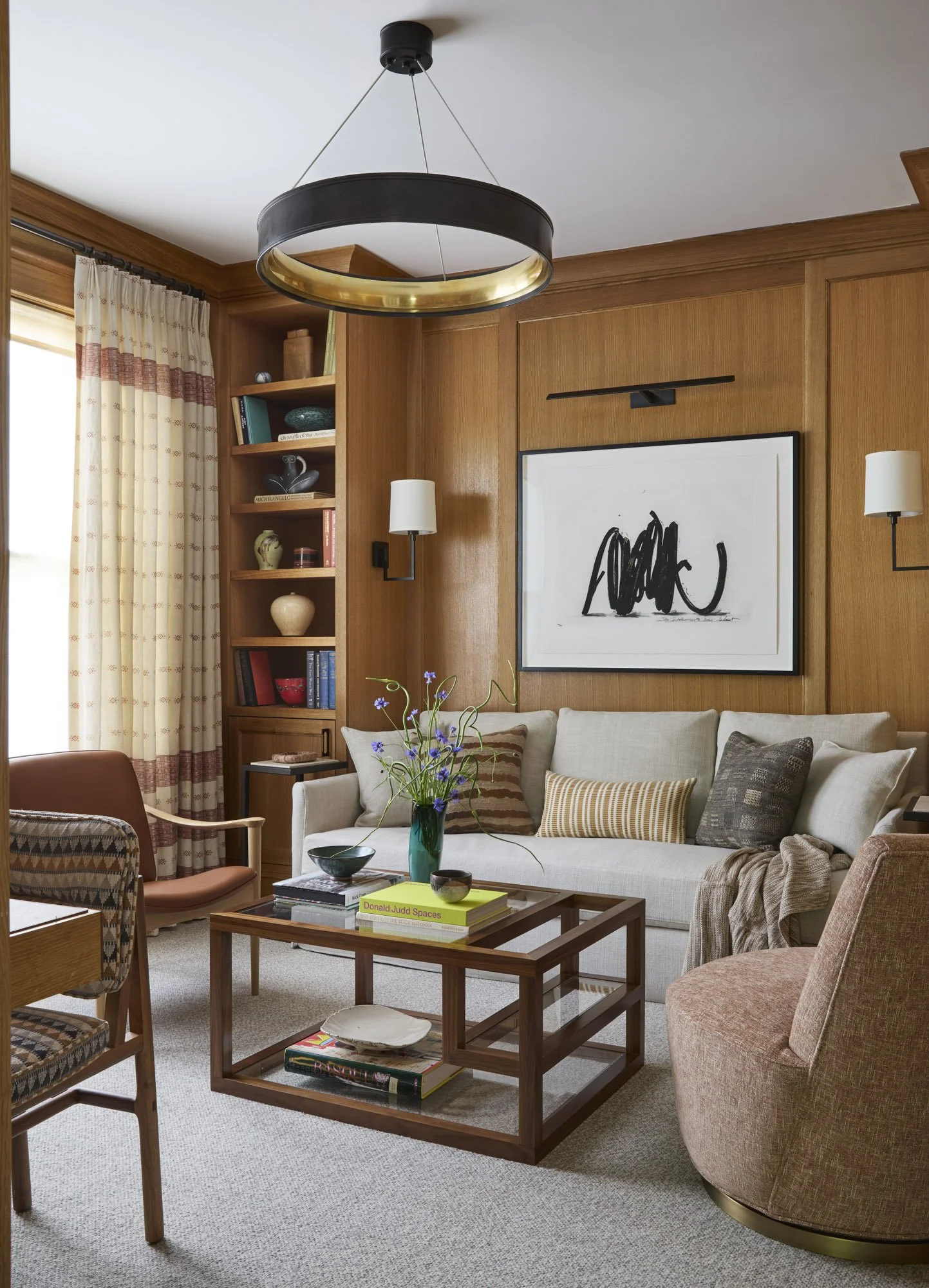 Person wearing a rabbit mask reads in a wood-paneled living room with abstract art, built-in shelving, and layered furnishings.