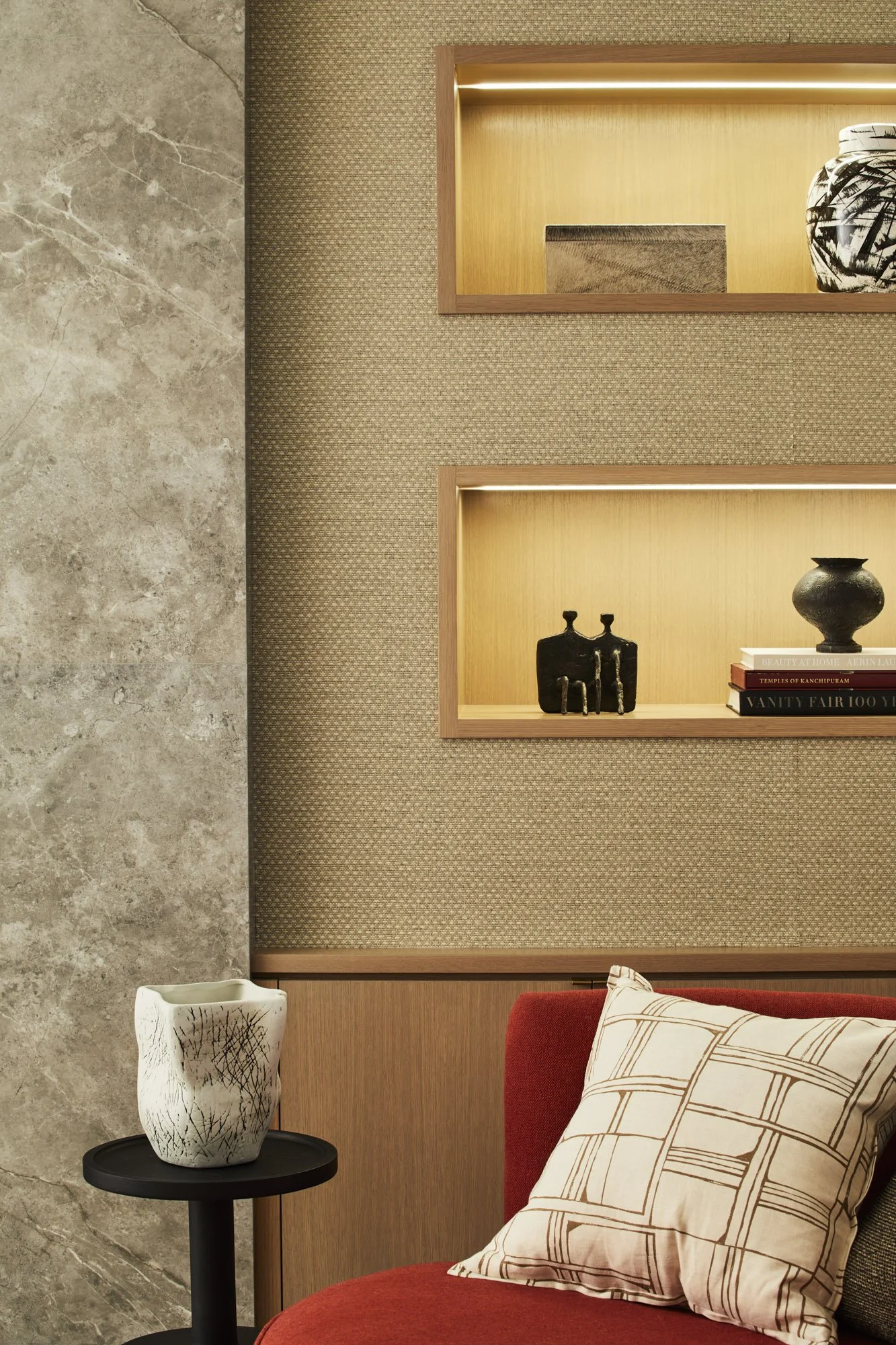 Red upholstered chair beside a textured wall and wood shelves styled with books, artwork, and decorative objects.