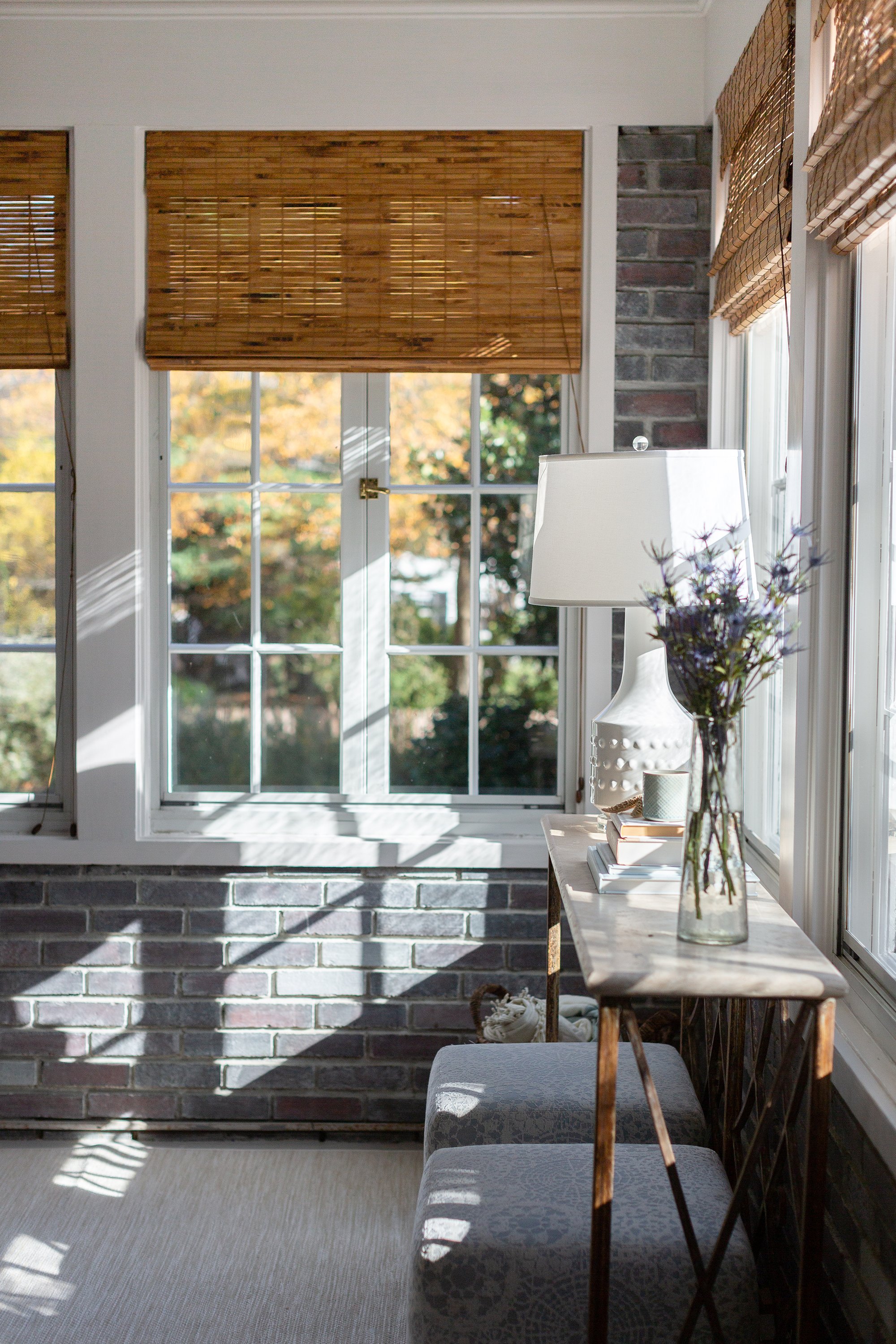Sunlit room with exposed brick walls, bamboo shades, upholstered stools, and a wood table styled with purple flowers and a white lamp.