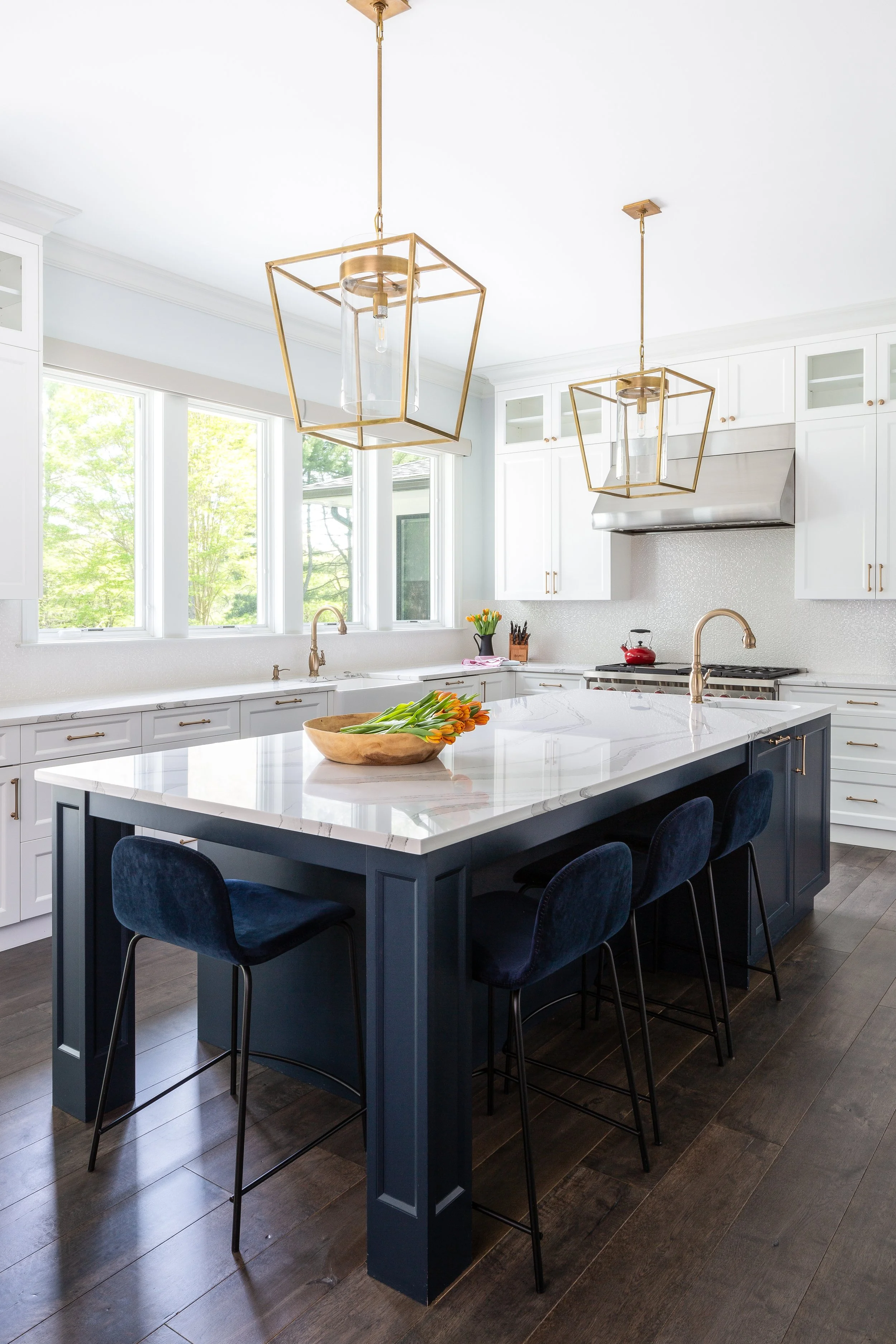 Regan arranging a chair in a white kitchen with a marble island, orange flowers, pendant lighting, and a stainless steel hood.
