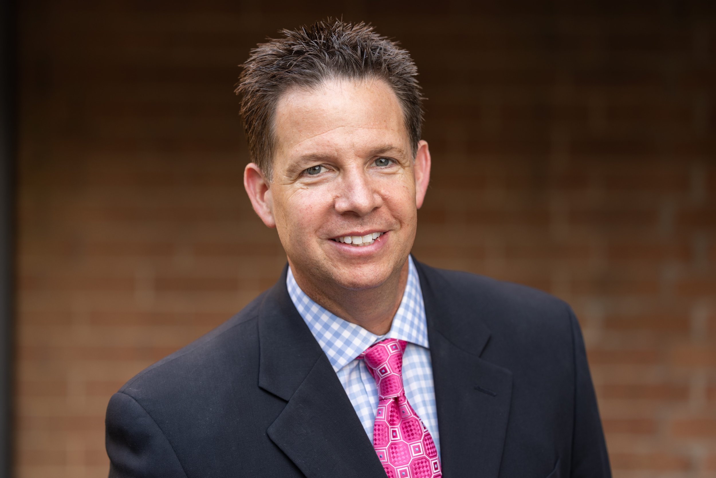 Wes Bowman smiling in a professional headshot wearing a dark suit jacket, blue checkered shirt, and pink tie.