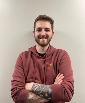 Young man with a beard and tattooed arm, smiling, wearing a maroon hoodie, standing with arms crossed against a plain wall.
