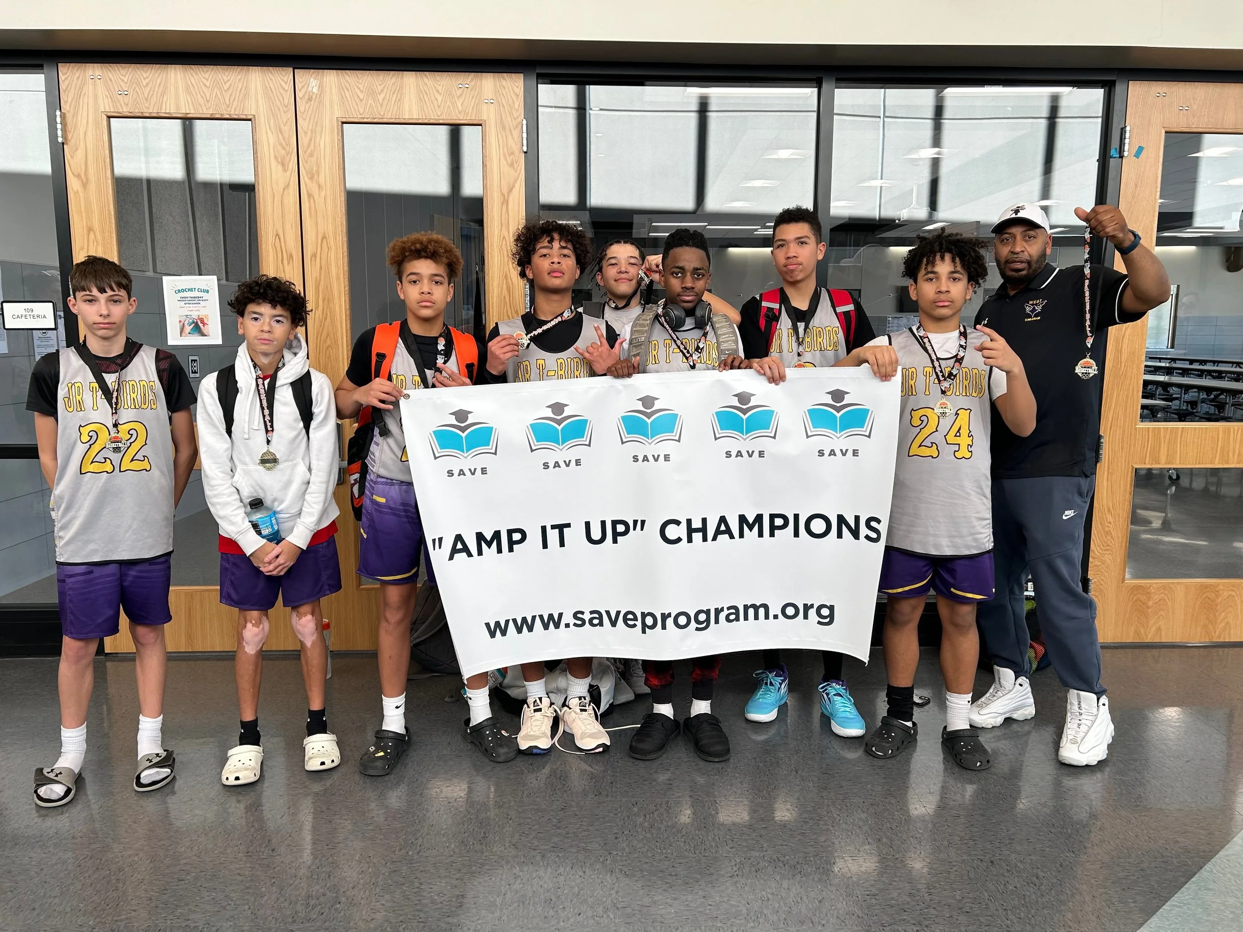 Group of young boys and their coach standing inside a school or community center, holding a large banner that reads "AMP IT UP" CHAMPIONS and includes a website link. The boys are wearing sports jerseys with numbers and medals around their necks, indicating they have won a competition.