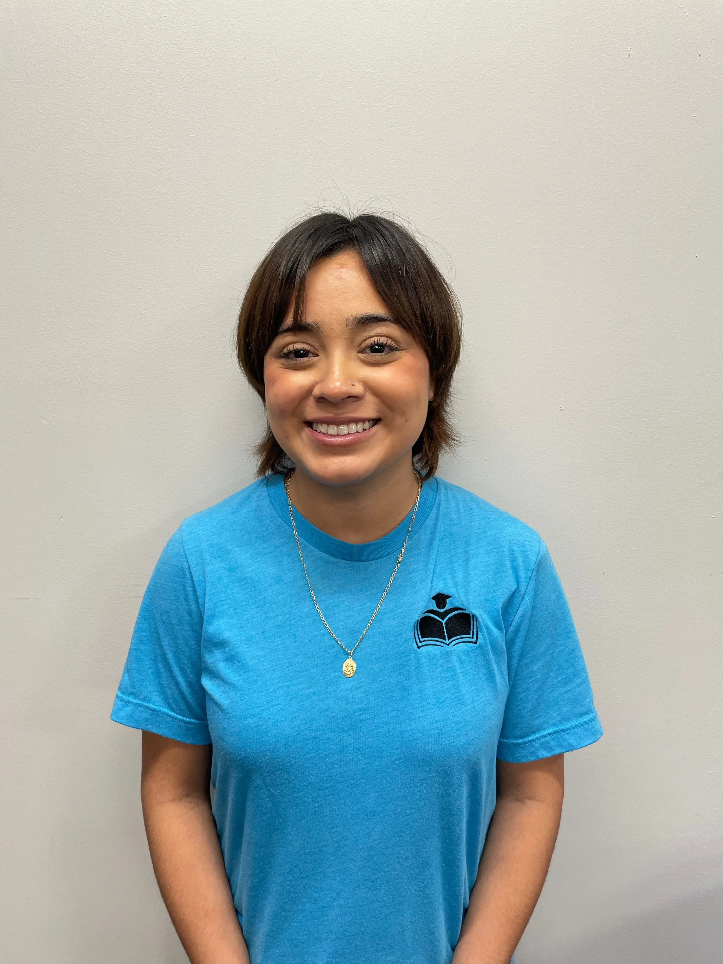 A young woman with short brown hair smiling at the camera, wearing a blue T-shirt with a logo of a graduation cap and book on it, standing against a plain light-colored wall.