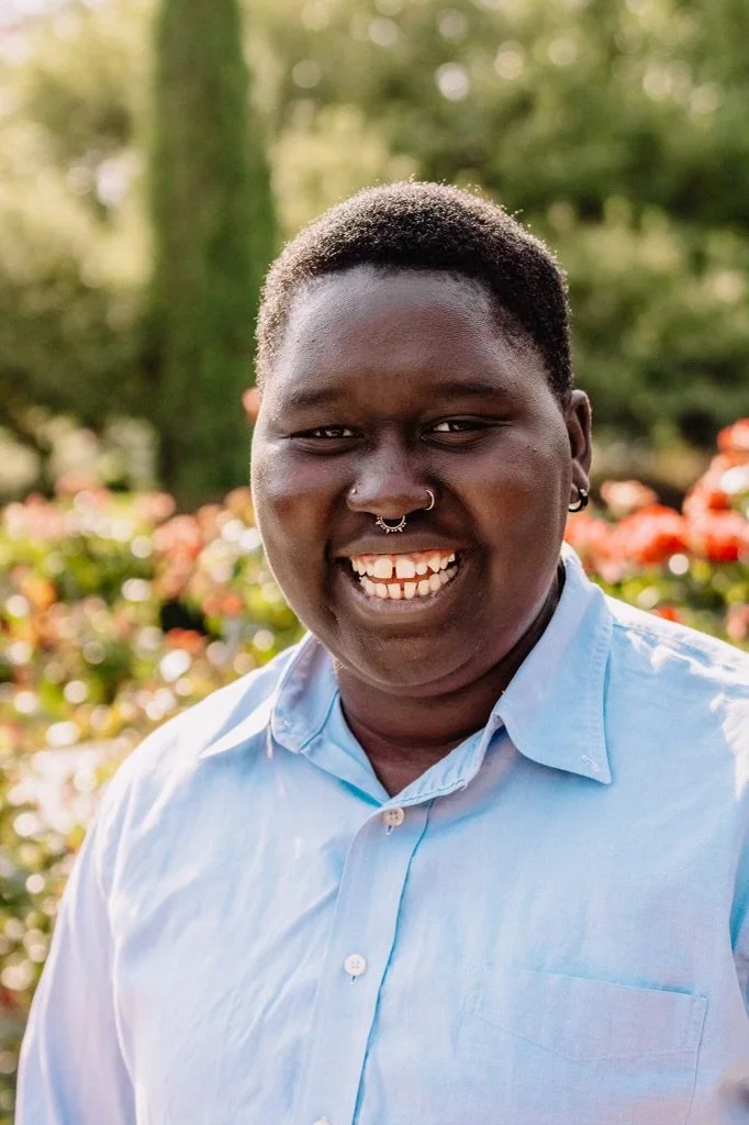 A person smiling outdoors in a garden with flowers in the background, wearing a light blue collared shirt.