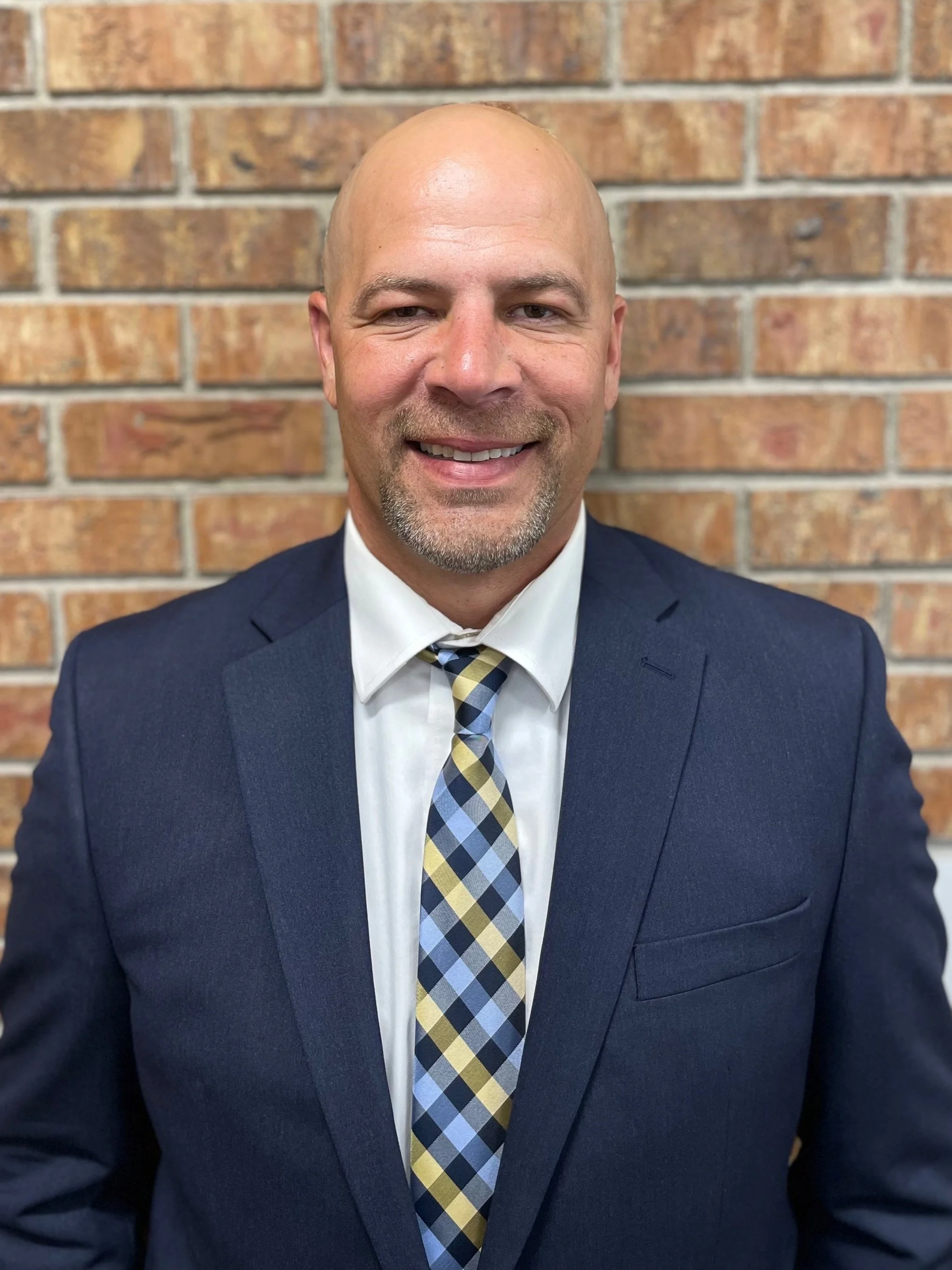 A man in a navy suit and a plaid tie, smiling, standing in front of a brick wall.