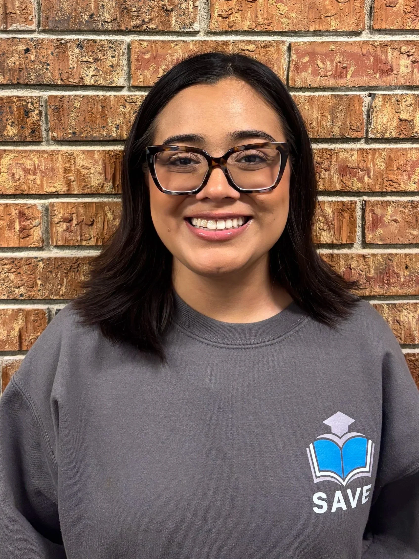 A young woman with short brown hair smiling at the camera, wearing a blue T-shirt with a logo of a graduation cap and book on it, standing against a plain light-colored wall.