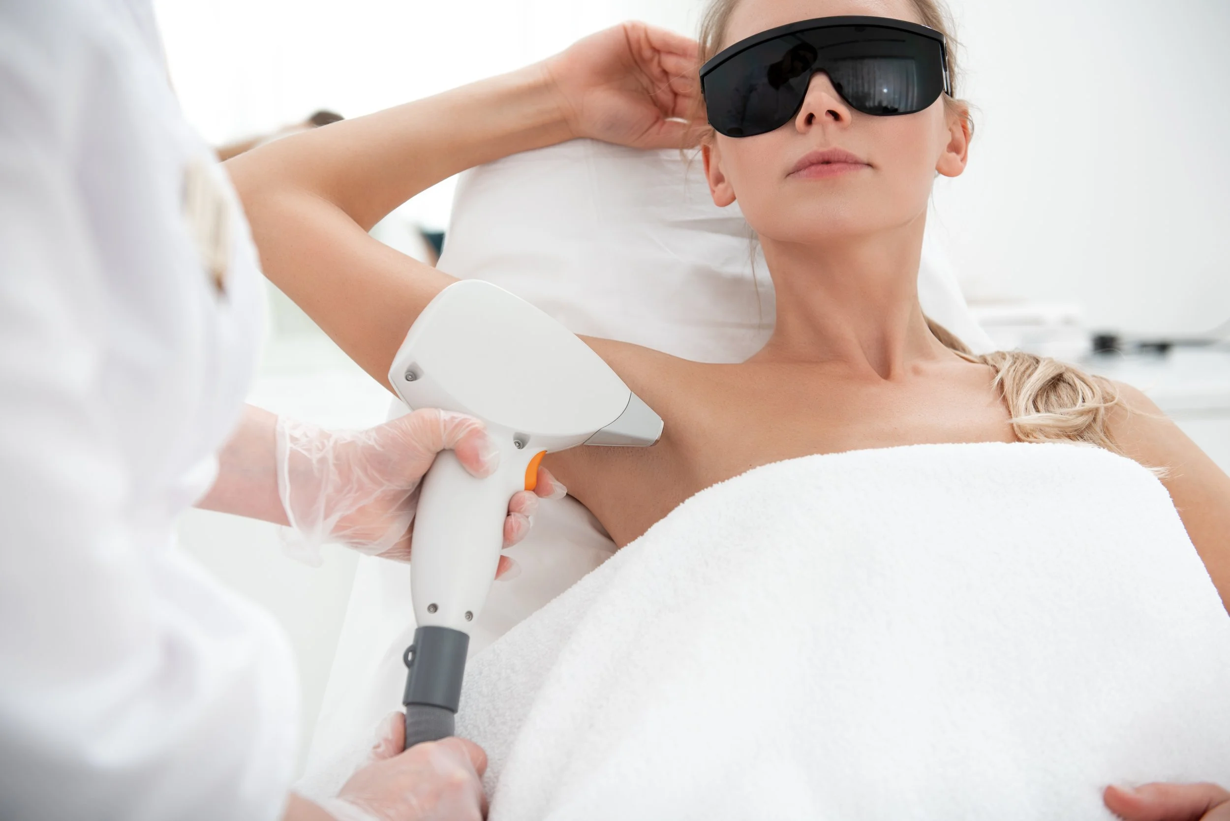 A woman getting a laser hair removal treatment on her underarm at a medical clinic, wearing protective eyewear.