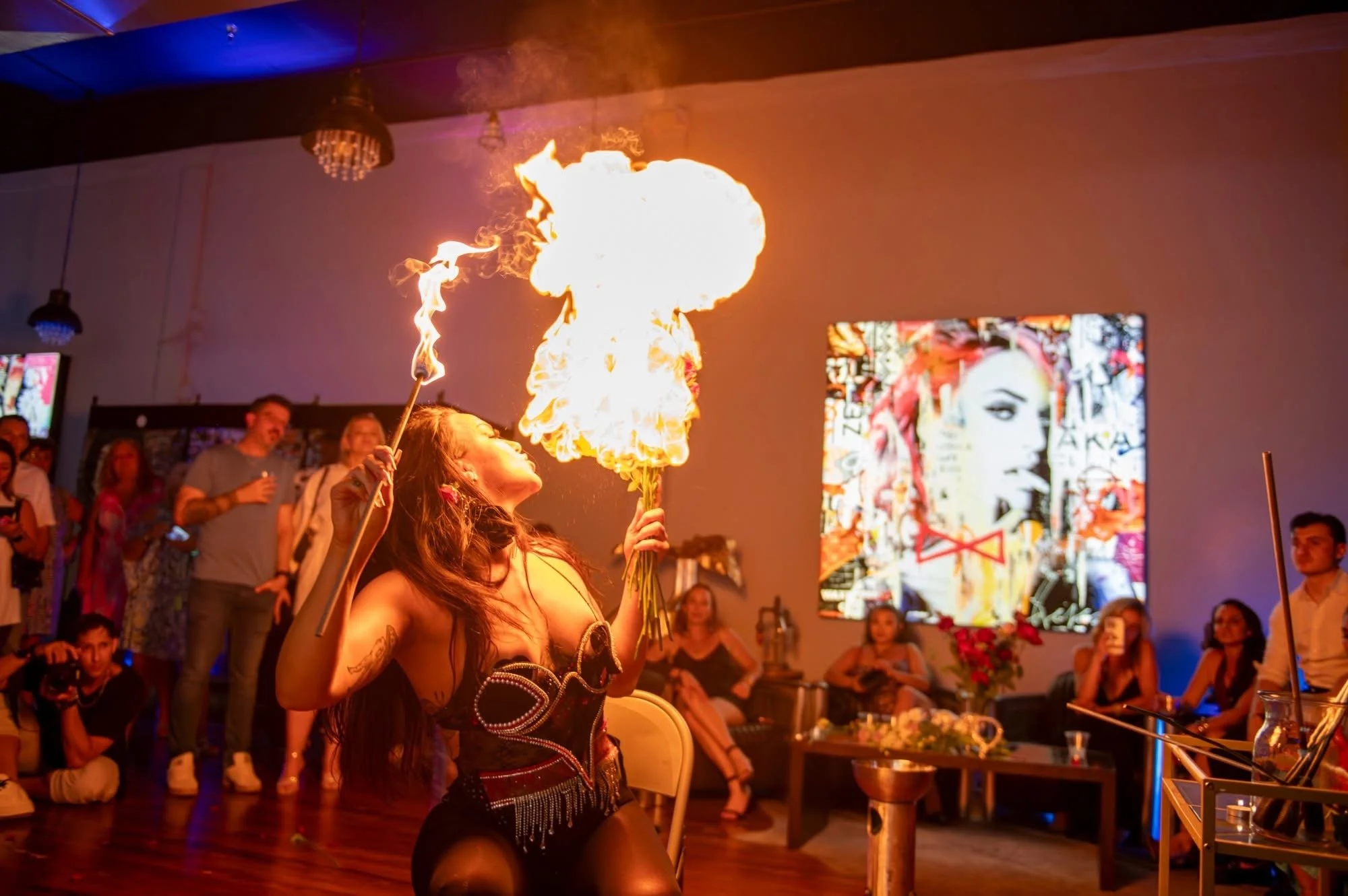 A woman performing a fire dance at an indoor event with an audience watching. She is holding fire torches, and flames are visible in the air.
