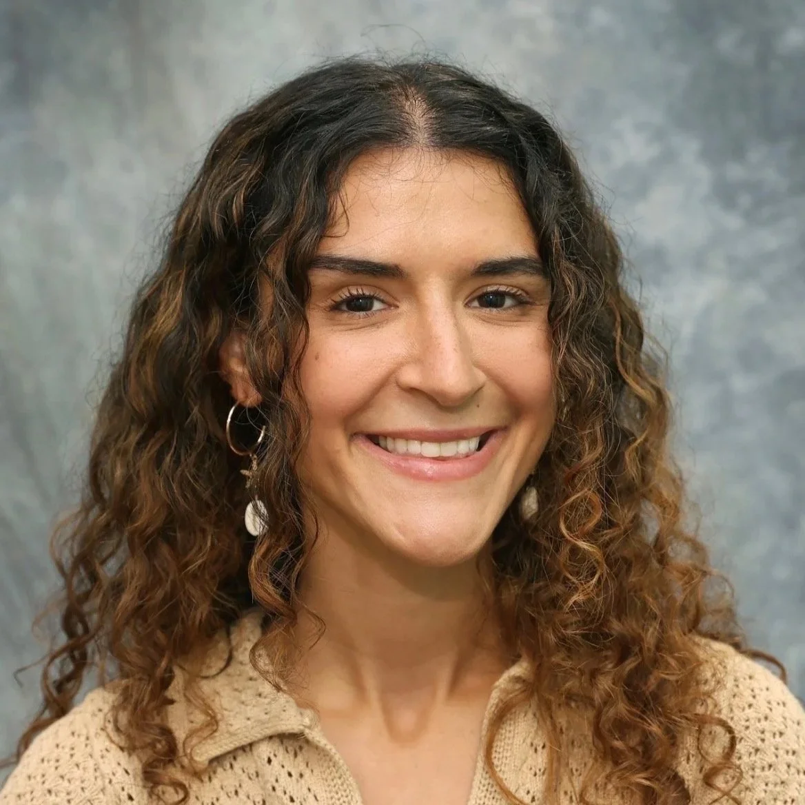 Smiling woman with curly hair, wearing a beige crochet top and hoop earrings, posed against a gray mottled background.