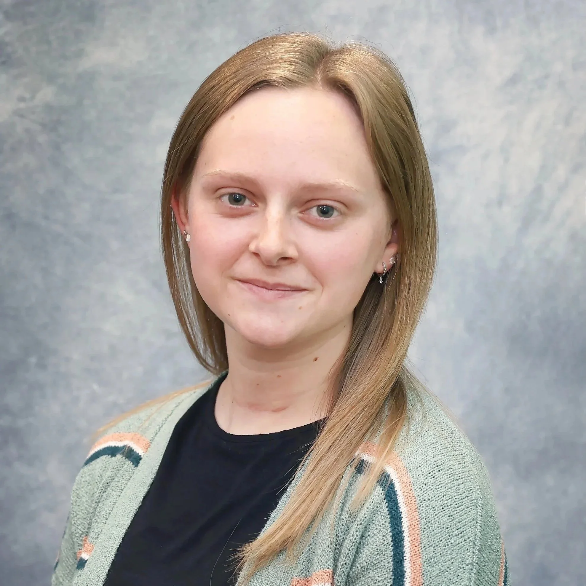 A young woman with shoulder-length light brown hair, wearing earrings, a black top, and a gray cardigan with orange, blue, and white stripes, standing against a gray textured background.