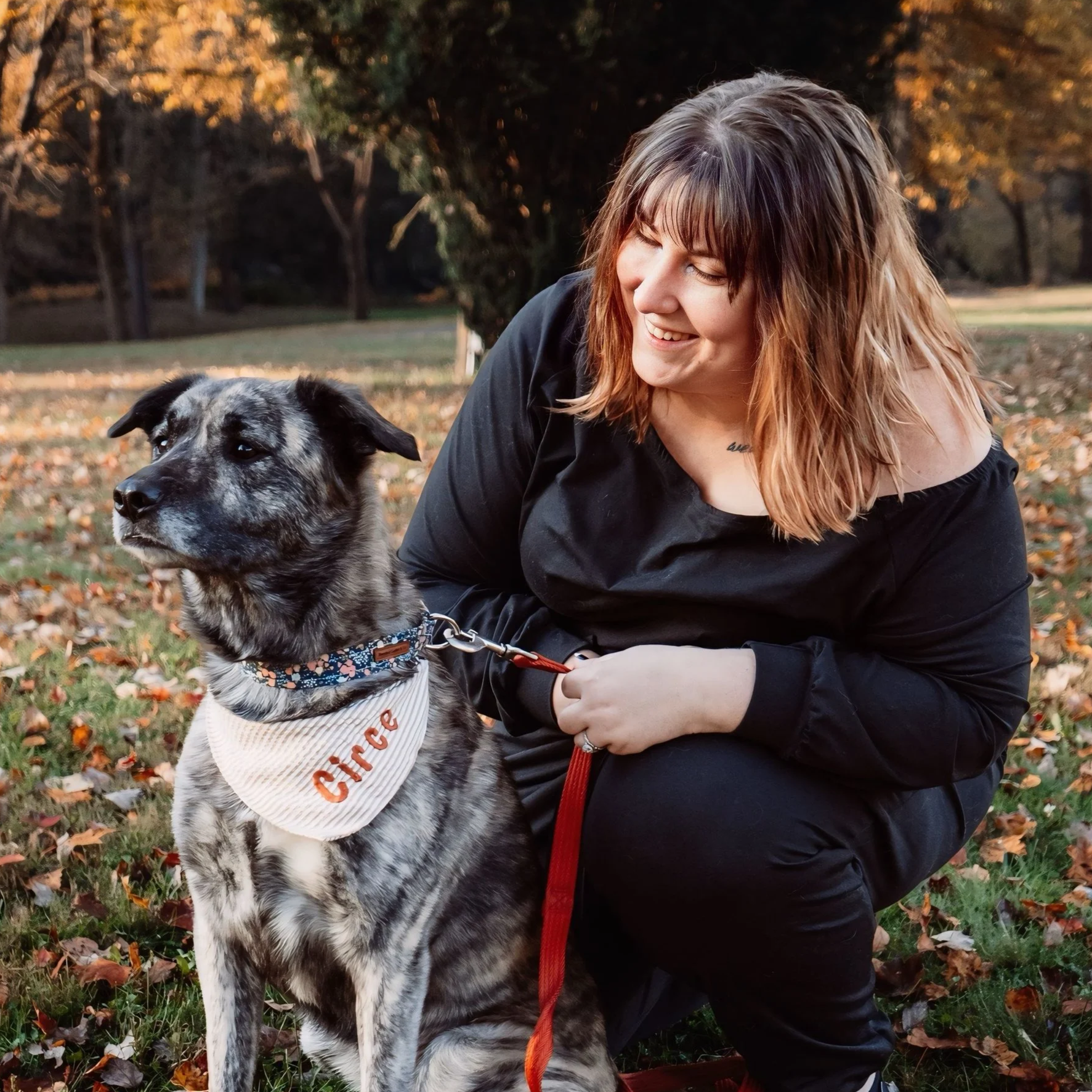 a smiling woman looking at a dog with a bandana inscribed with "circe" they are surrounded by fall leaves
