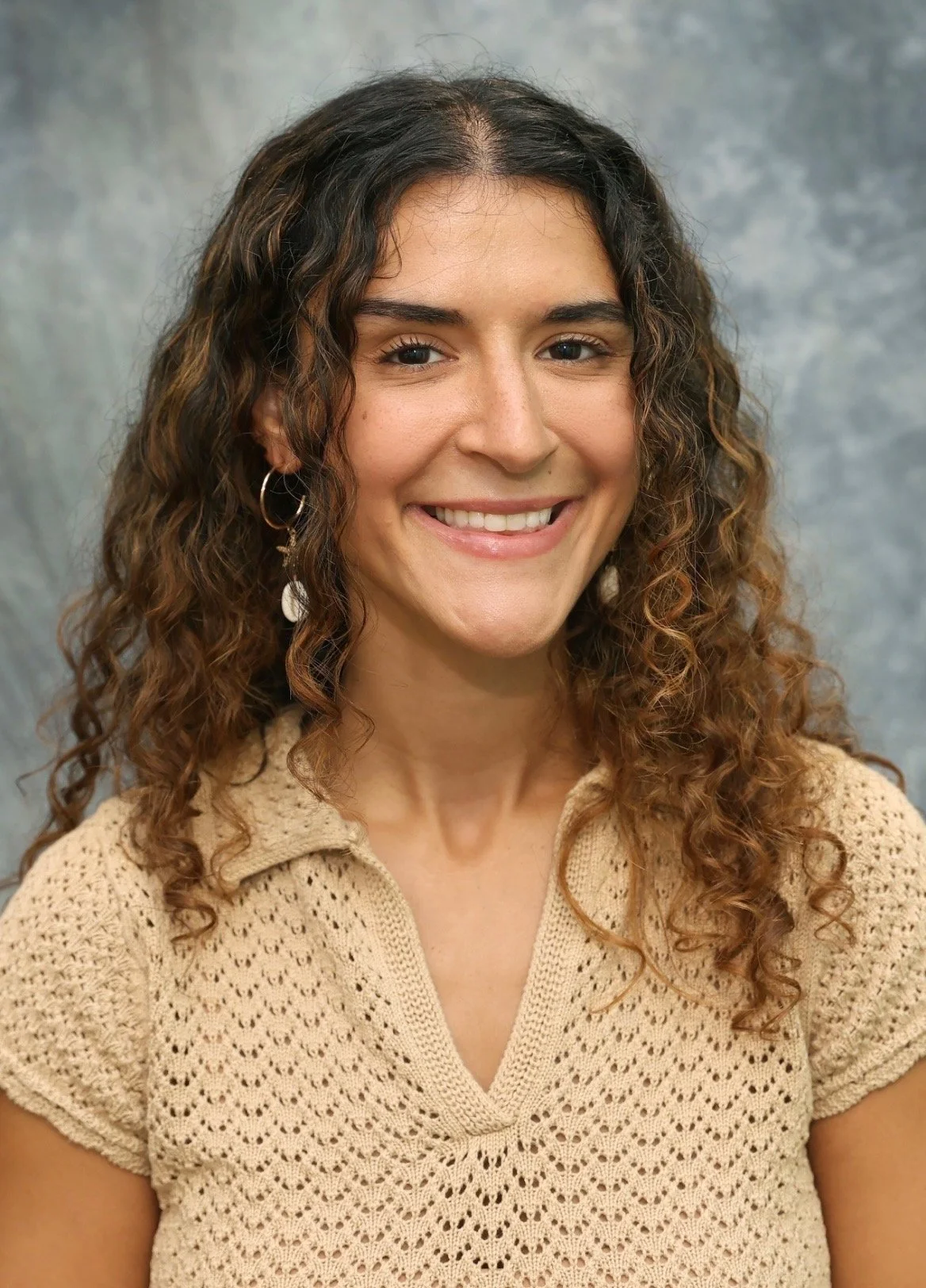 Smiling woman with curly hair, wearing a beige crochet top and hoop earrings, posed against a gray mottled background.