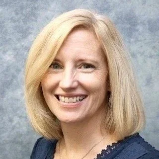 Blond woman smiling with a necklace, wearing a navy blouse.