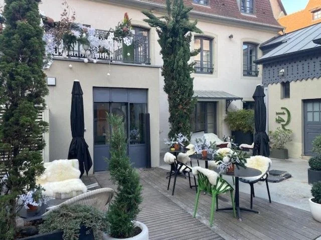 Outdoor patio with table and chairs, surrounded by potted plants, umbrellas, and a building with windows and balcony