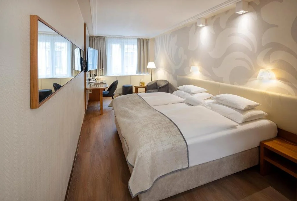 Hotel room with a large bed, wall-mounted TV, seating area with armchair and table, windows with curtains, and warm lighting.