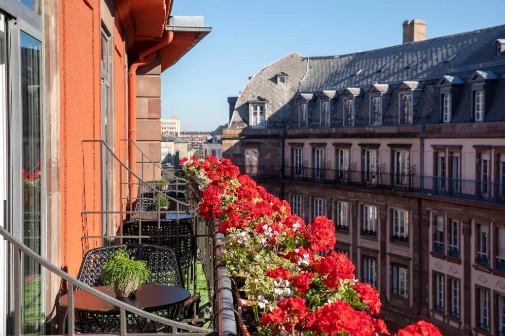 Balcony with black metal chairs, potted plants, and a railing filled with vibrant red and white flowers, overlooking classic European-style buildings in a cityscape under a clear blue sky.