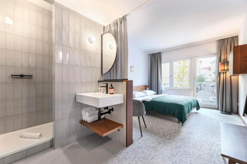A modern hotel room with a bed, large window, gray curtains, a small desk, and a bathroom area with a sink and shower.