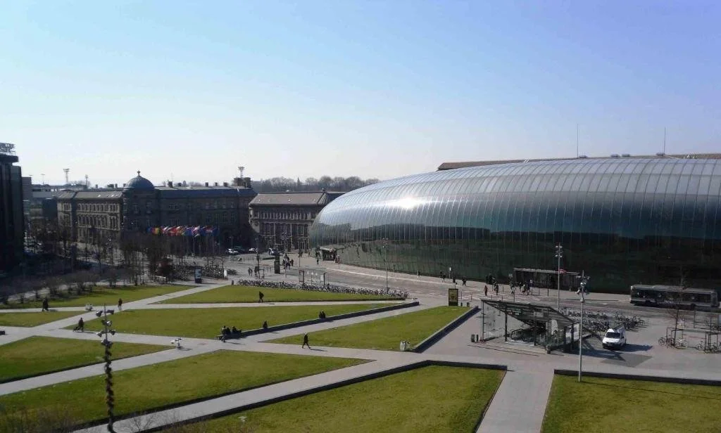 A modern glass building with a rounded roof next to a park with green lawns and walking paths, with people walking and a bus parked nearby.