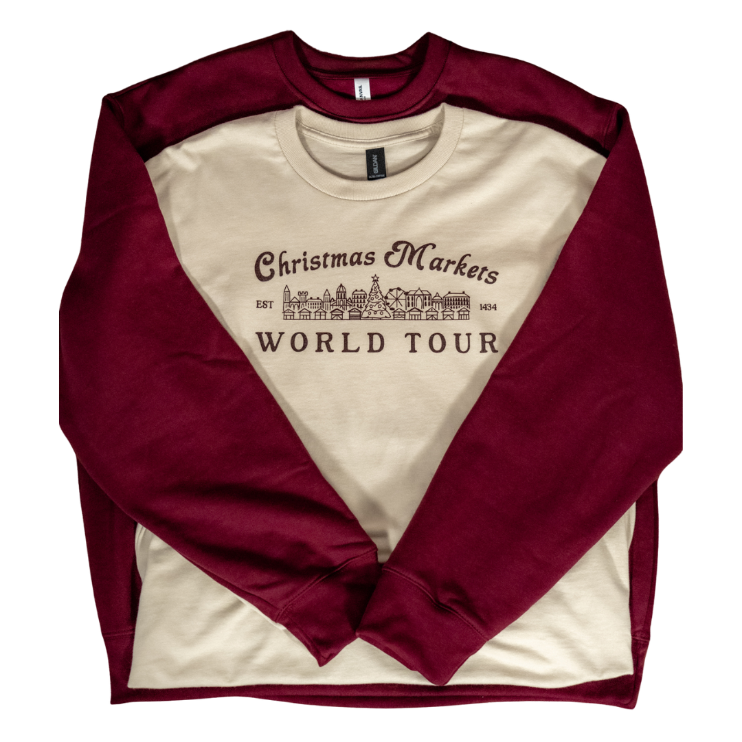 Two long-sleeve shirts, one cream with maroon sleeves, featuring text 'Christmas Markets World Tour' and a line drawing of Christmas market stalls and a tree.