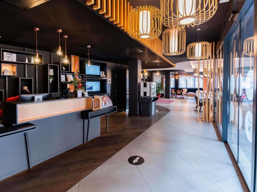 Modern hotel lobby with a reception desk, decorative lighting, wooden ceiling accents, and a sitting area with chairs and large windows.