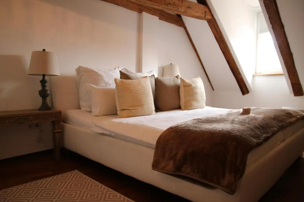 A cozy bedroom with a bed, multiple pillows, a blanket, and wooden beams on the ceiling, lit by natural light from a window.