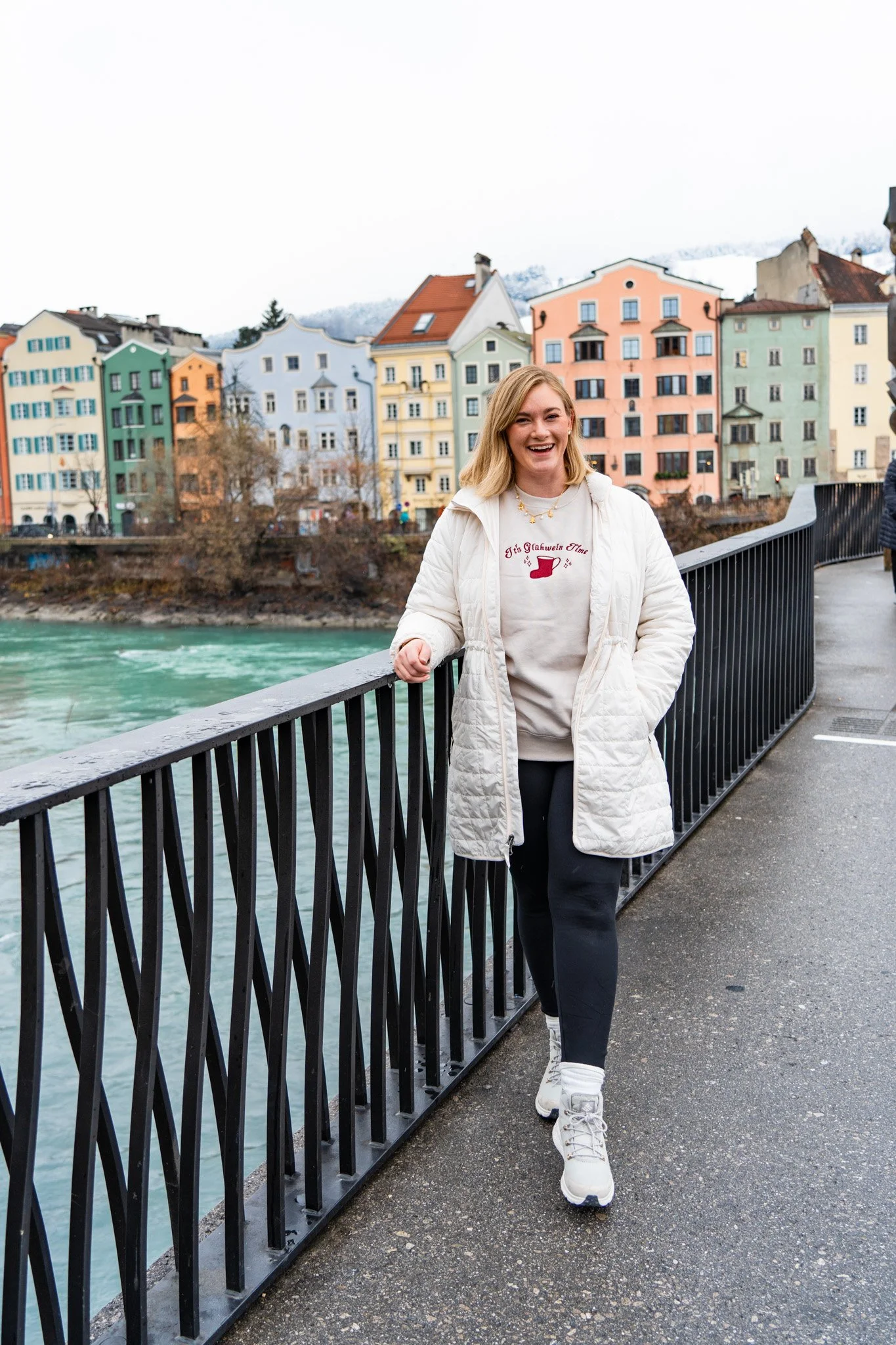 A woman in a white jacket, black pants, and white sneakers walking on a bridge with a black railing. She is smiling and has blond hair. Behind her are colorful buildings and a river, with a mountain visible in the background.