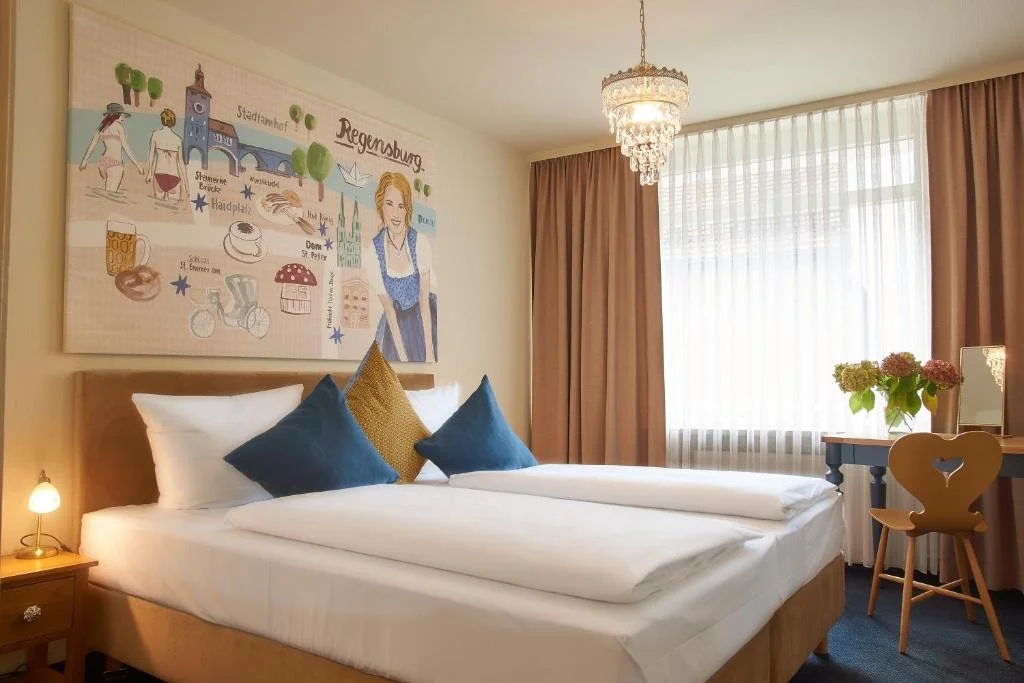 A bedroom with a wooden bed, white bedding, and three pillows, two blue and one gold. There is a nightstand with a small lamp on the left side. The wall behind the bed has a large colorful illustration featuring landmarks, people, and scenes from Regensburg, Germany. A chandelier hangs from the ceiling. To the right, there's a window with sheer white curtains and pink drapes, a vase with pink and green flowers, a mirror, and a wooden desk with a heart-shaped chair.