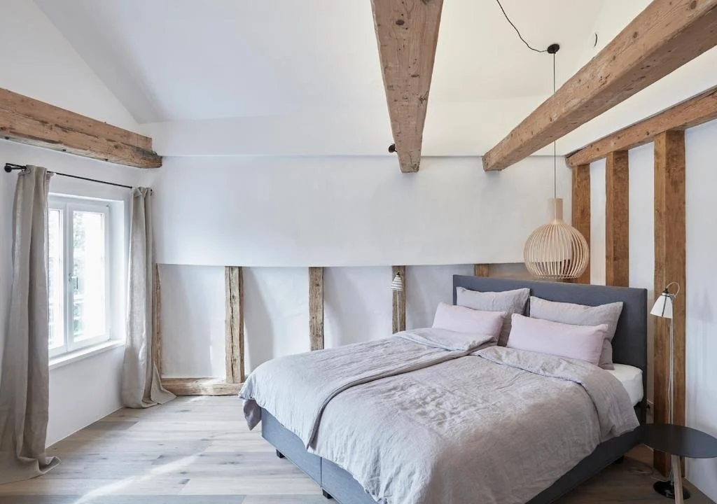 A modern bedroom with a sloped ceiling, featuring wooden beams and a large window with beige curtains. The bed has a grey headboard and light-colored bedding, with a round hanging light fixture and a small black bedside table.