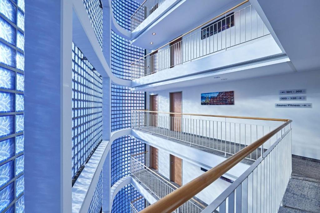 Interior view of a modern hotel or office building with multiple floors, white railings, glass-block windows, and blue accent lighting. There are doors on each floor and signs indicating directions to different rooms and amenities.