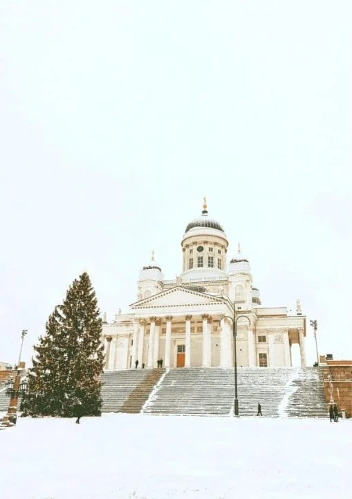 A white historic building with columns and a dome, snow-covered steps, and trees, under a cloudy sky.