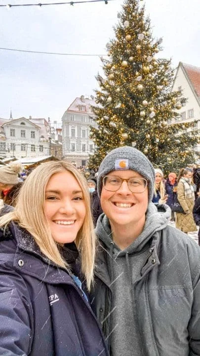 A smiling woman and man taking a selfie in front of a lit Christmas tree in a snowy outdoor town square with people in winter clothing and historic buildings in the background.