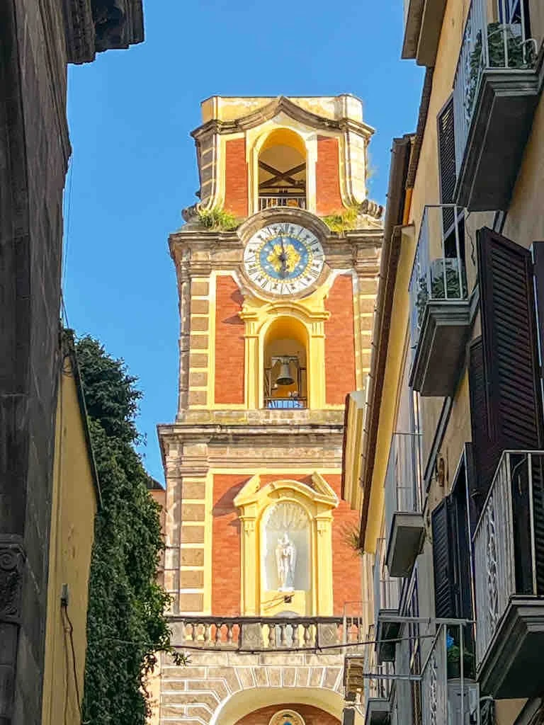 A tall historic clock tower with a bright blue sky in the background, surrounded by buildings with balconies.