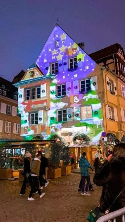 Building decorated with colorful lights and Christmas-themed projections, in a festive outdoor setting with people walking around at night.