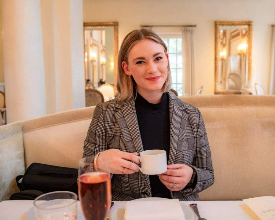 A woman with blonde hair wearing a plaid blazer and black top, sitting at a table in a well-lit restaurant, holding a white cup, smiling at the camera.