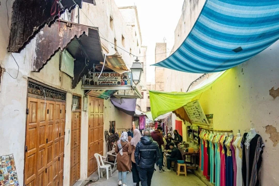 People walking and shopping in a narrow outdoor market alley with colorful fabric canopies and clothing displays on the right side.