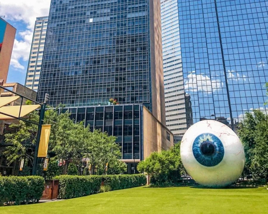 Large artificial eyeball sculpture in a city park, surrounded by greenery and high-rise office buildings with reflective glass facades.