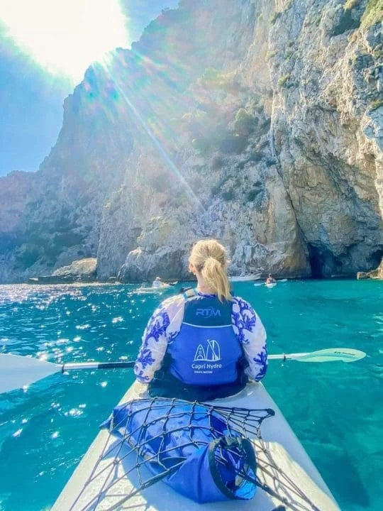 A woman kayaking on clear blue water with a rocky cliff and bright sunlight in the background.