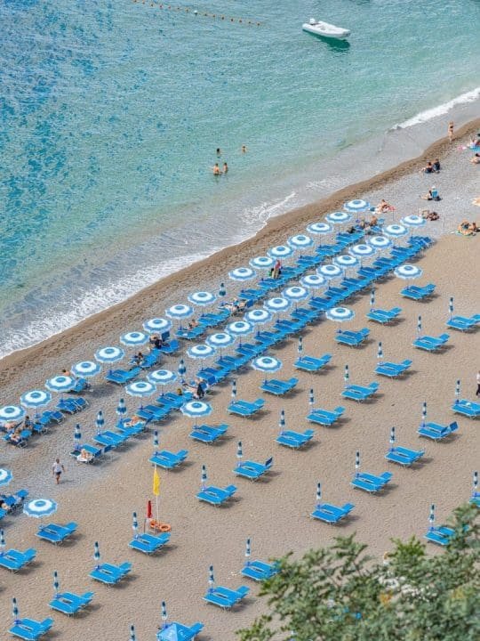 Empty blue beach chairs and umbrellas arranged in rows on sandy beach near ocean, with a few people swimming and a boat in the water.