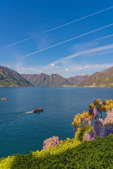 A lake surrounded by mountains with colorful flowers and bushes in the foreground. A boat is on the water, and the sky has some clouds and airplane contrails.