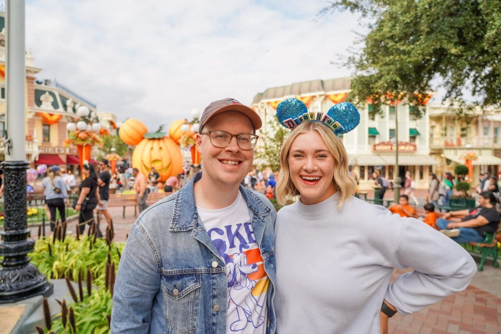 A smiling man and woman posing together at a theme park decorated for Halloween, with a large carved pumpkin figure in the background, wearing Disneyland-themed headbands.