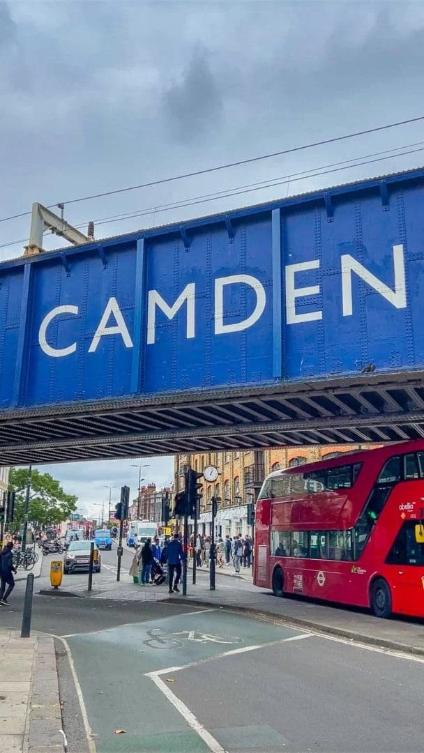 Street scene in Camden with a blue bridge overhead bearing the word 'Camden', a red double-decker bus, pedestrians crossing, and a bike lane.